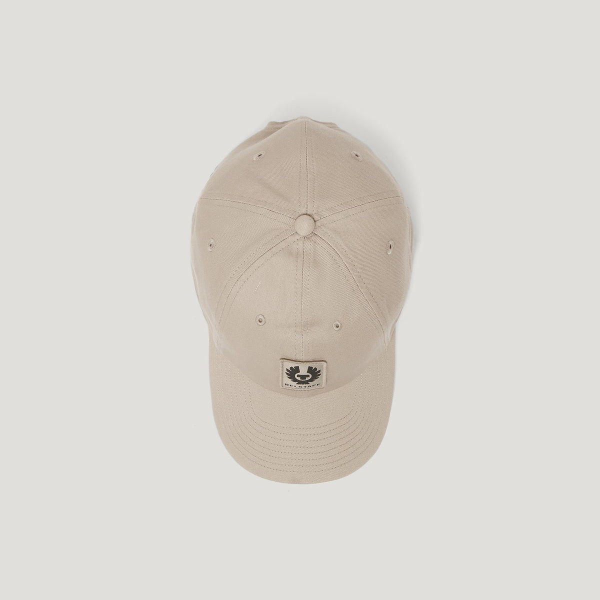 A light beige baseball cap with a small black embroidered patch on the front of the brim, shown against a solid light gray background. Belstaff Cap. Sakks Menswear Southampton.