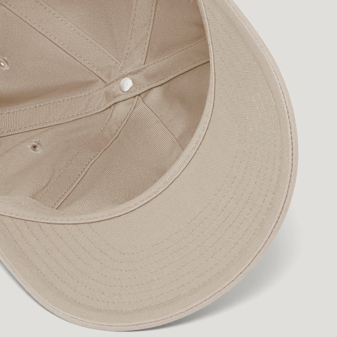 Close-up detail shot of a beige baseball cap showing the curved brim, stitched panel seams, and a single white snap button on a light gray background. Belstaff Cap. Sakks Menswear Southampton.