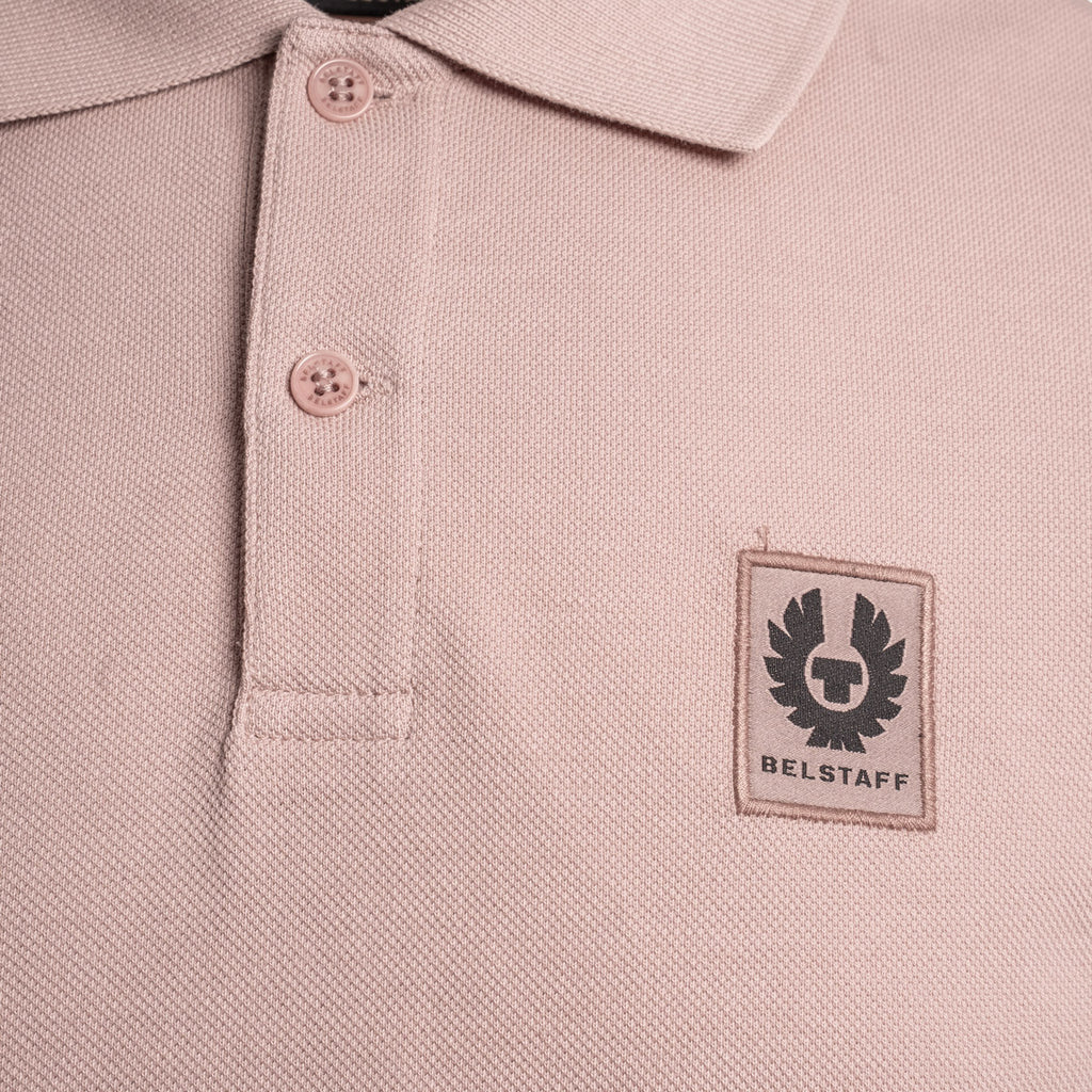 Close-up of a light pink textured polo shirt with a black Belstaff winged logo patch on the left chest. Belstaff Polo. Sakks Menswear Southampton.