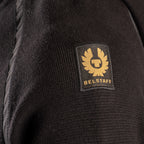 Close-up of a black ribbed knit garment featuring a square black patch with a gold BELSTAFF logo and text. Belstaff Hoodie. Sakks Menswear Southampton.