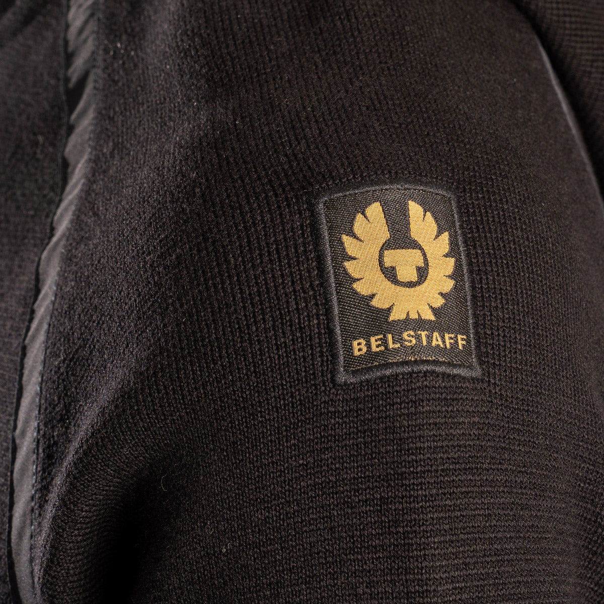Close-up of a black ribbed knit garment featuring a square black patch with a gold BELSTAFF logo and text. Belstaff Hoodie. Sakks Menswear Southampton.