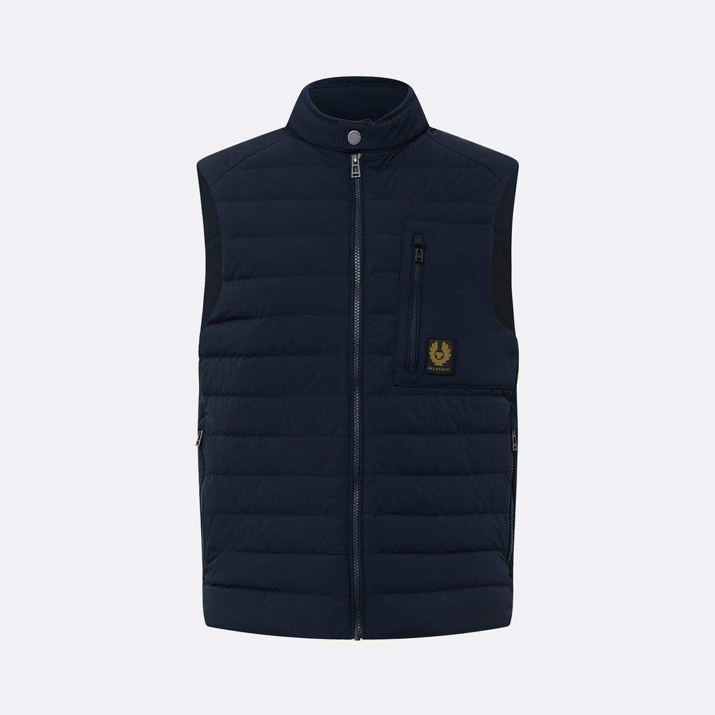 A dark navy blue quilted puffer vest with a stand-up collar, front zipper, and a vertical zippered chest pocket featuring a small yellow embroidered logo, shown against a light gray background. Belstaff Gilet. Sakks Menswear Southampton.