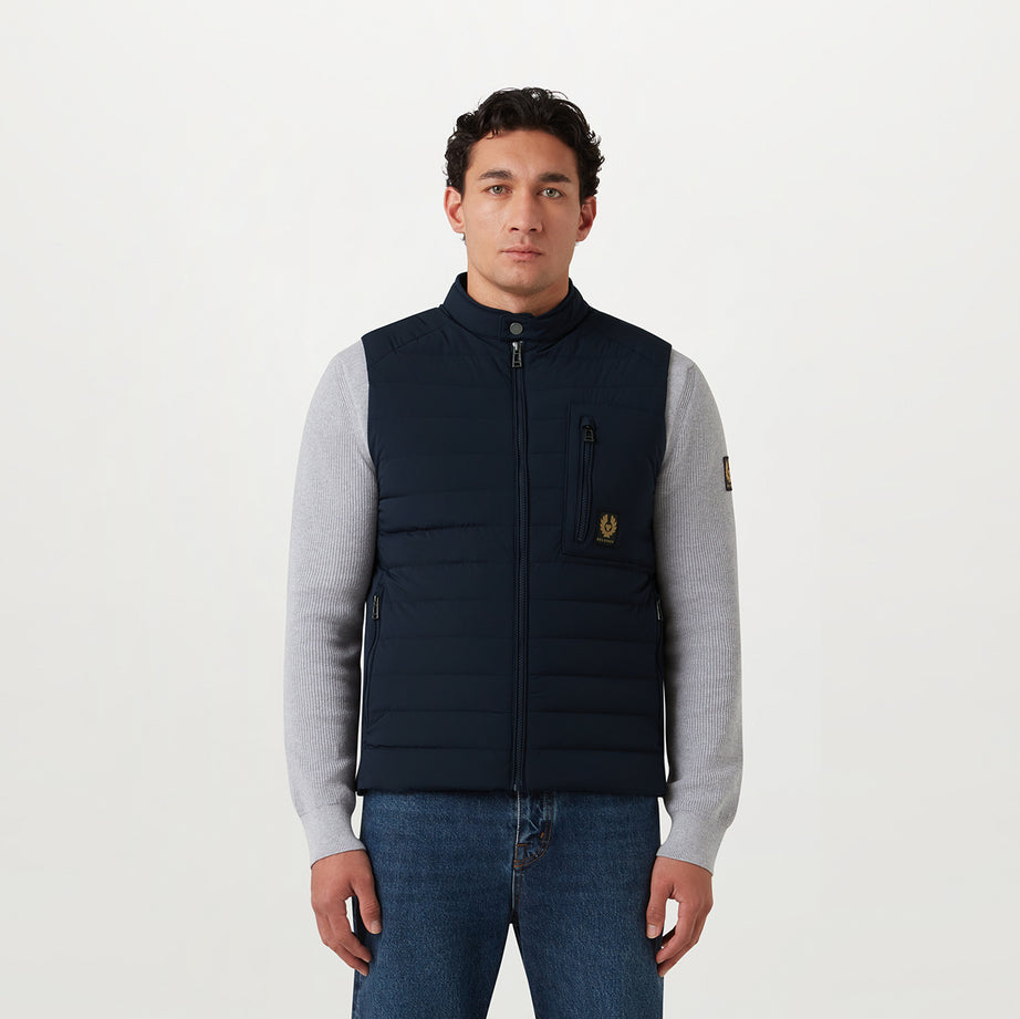 A man wearing a navy blue puffer vest over a light gray long-sleeve shirt and blue jeans, standing against a plain white studio background. Belstaff Gilet. Sakks Menswear Southampton.