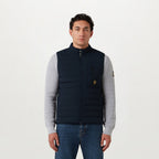 A man wearing a navy blue puffer vest over a light gray long-sleeve shirt and blue jeans, standing against a plain white studio background. Belstaff Gilet. Sakks Menswear Southampton.