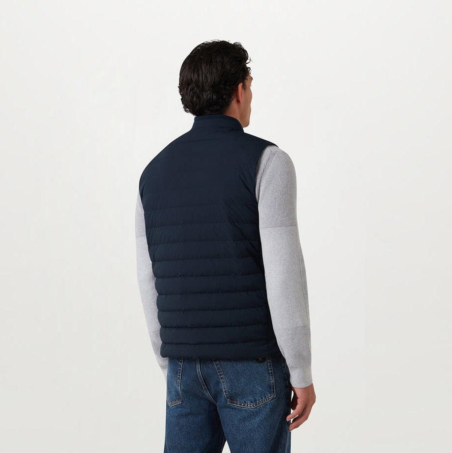 A man shown from the back wearing a dark navy quilted puffer vest over a light gray long-sleeve shirt and blue jeans, against a plain white studio background. Belstaff Gilet. Sakks Menswear Southampton.