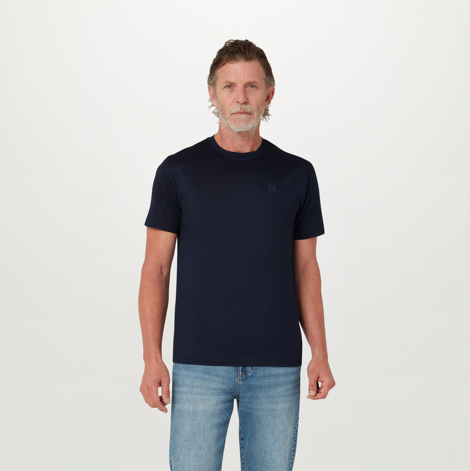 A man wearing a dark navy blue crew-neck t-shirt with a small tonal embroidered logo on the left chest, paired with light-wash blue jeans, standing against a plain white studio background. Belstaff T-Shirt. Sakks Menswear Southampton.