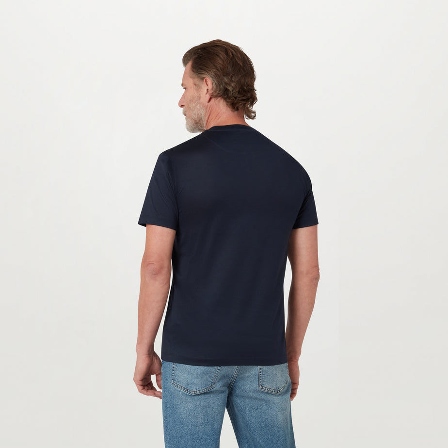 A man viewed from the back wearing a dark navy blue short-sleeve crewneck t-shirt and light blue jeans, standing against a plain white studio background. Belstaff T-Shirt. Sakks Menswear Southampton.