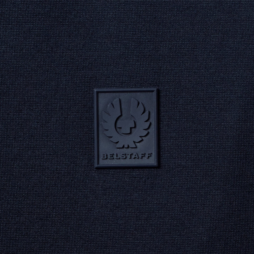 A close-up of a dark navy blue textured fabric with a square black rubber patch featuring the embossed BELSTAFF logo and brand name. Belstaff T-Shirt. Sakks Menswear Southampton.