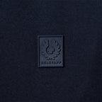A close-up of a dark navy blue textured fabric with a square black rubber patch featuring the embossed BELSTAFF logo and brand name. Belstaff T-Shirt. Sakks Menswear Southampton.