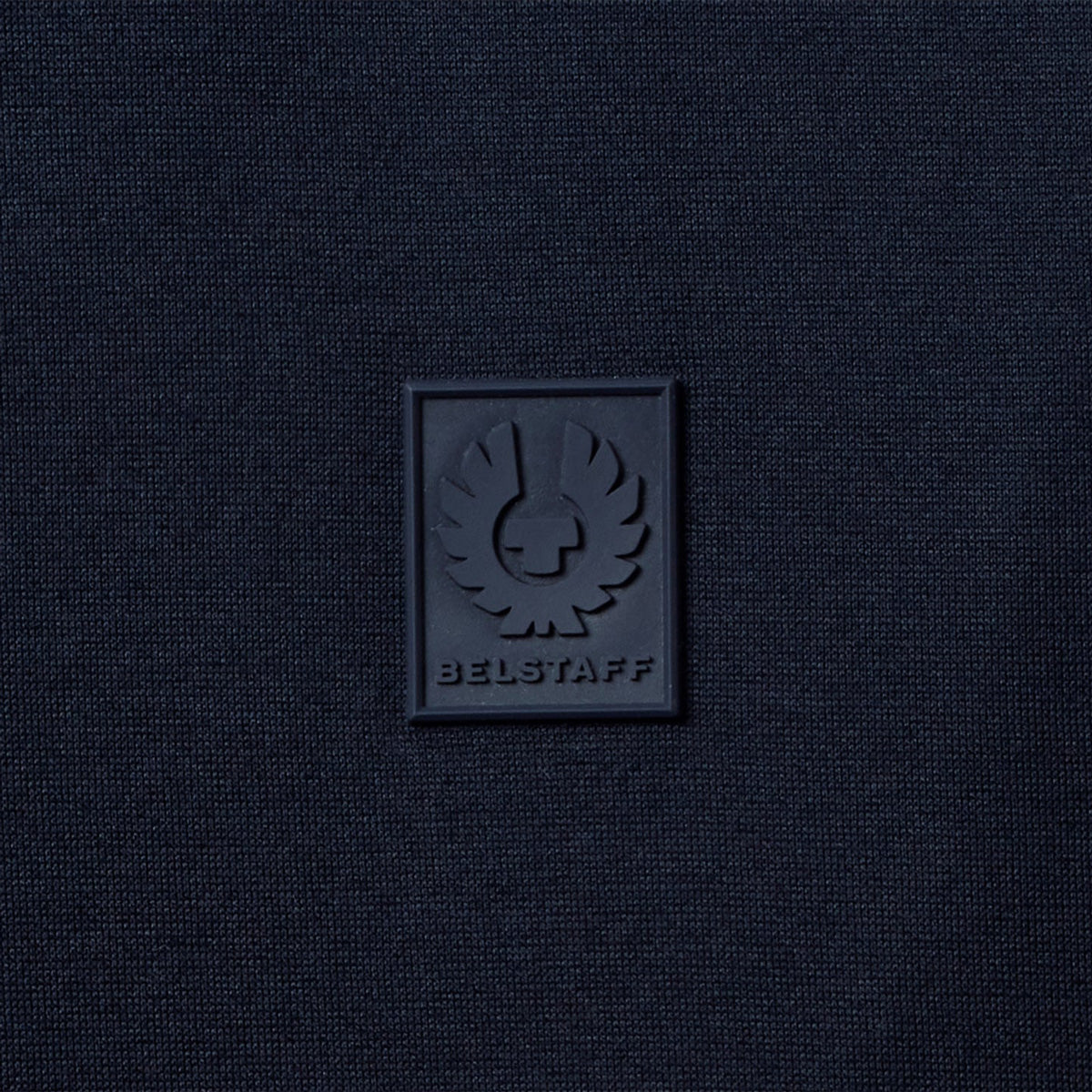 A close-up of a dark navy blue textured fabric with a square black rubber patch featuring the embossed BELSTAFF logo and brand name. Belstaff T-Shirt. Sakks Menswear Southampton.