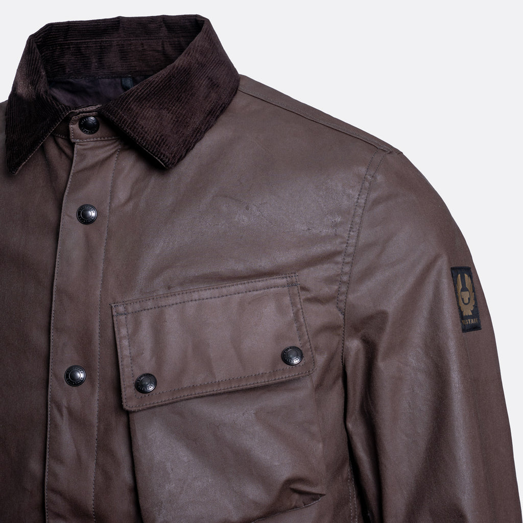 Close-up of a brown waxed cotton jacket with a dark corduroy collar, snap-button front, and a Brixton logo patch on the sleeve. Belstaff Jacket. Sakks Menswear Southampton.