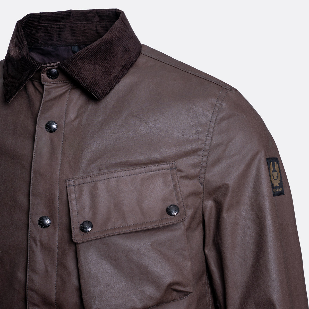 Close-up of a brown waxed cotton jacket with a dark corduroy collar, snap-button front, and a Brixton logo patch on the sleeve. Belstaff Jacket. Sakks Menswear Southampton.
