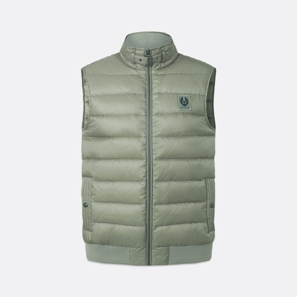 A sleeveless quilted puffer vest in a muted sage green color, featuring a stand-up collar, front zipper, and a small circular logo patch on the left chest, shown against a light gray background. Belstaff Gilet. Sakks Menswear Southampton.