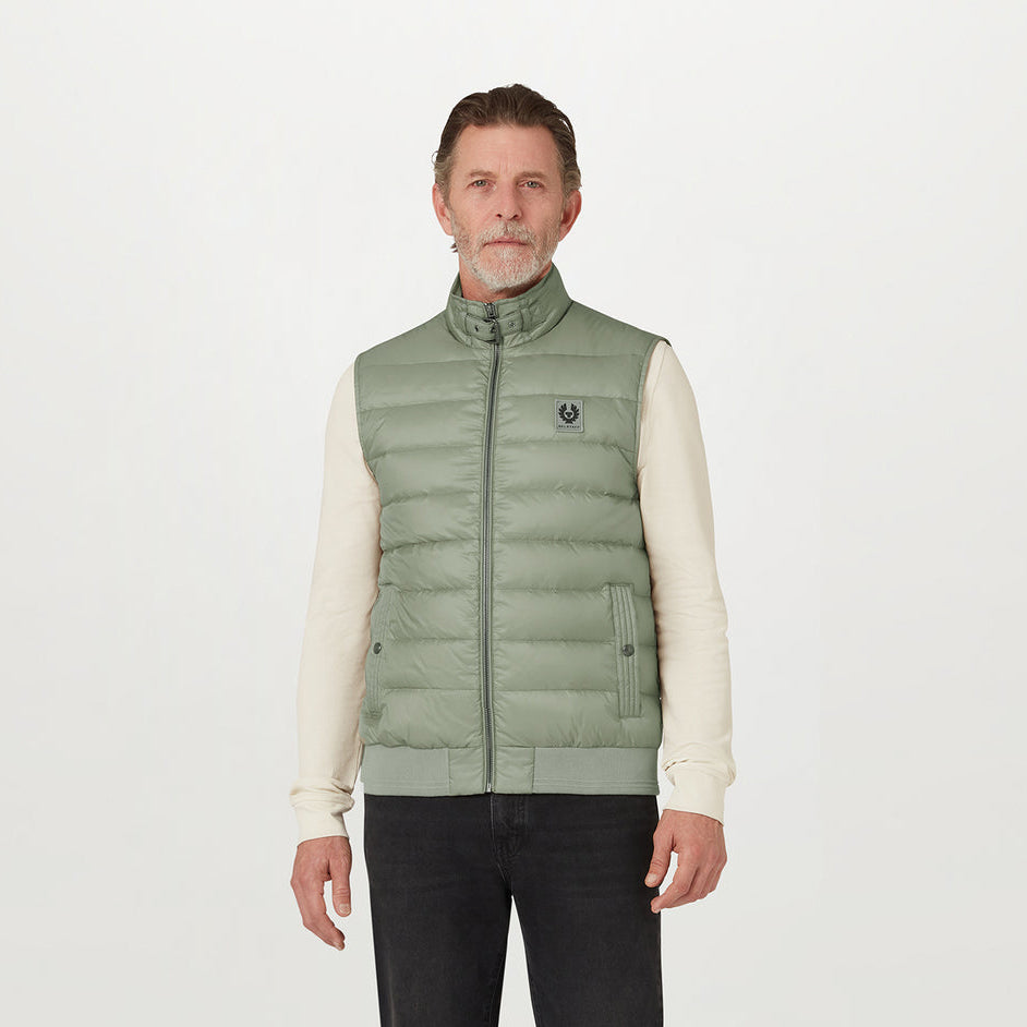 A man wearing a light sage green puffer vest over a cream long-sleeve shirt and black pants, standing against a plain white studio background. The vest features a small embroidered logo on the left chest. Belstaff Gilet. Sakks Menswear Southampton.
