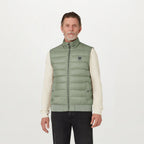 A man wearing a light sage green puffer vest over a cream long-sleeve shirt and black pants, standing against a plain white studio background. The vest features a small embroidered logo on the left chest. Belstaff Gilet. Sakks Menswear Southampton.