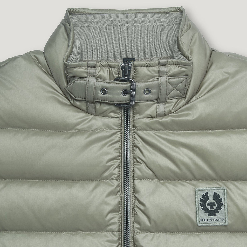 Close-up of a puffer jacket in a muted sage green color, featuring a high collar with a buttoned strap detail, a front zipper, and a square Belstaff logo patch on the lower right side. Belstaff Gilet. Sakks Menswear Southampton.