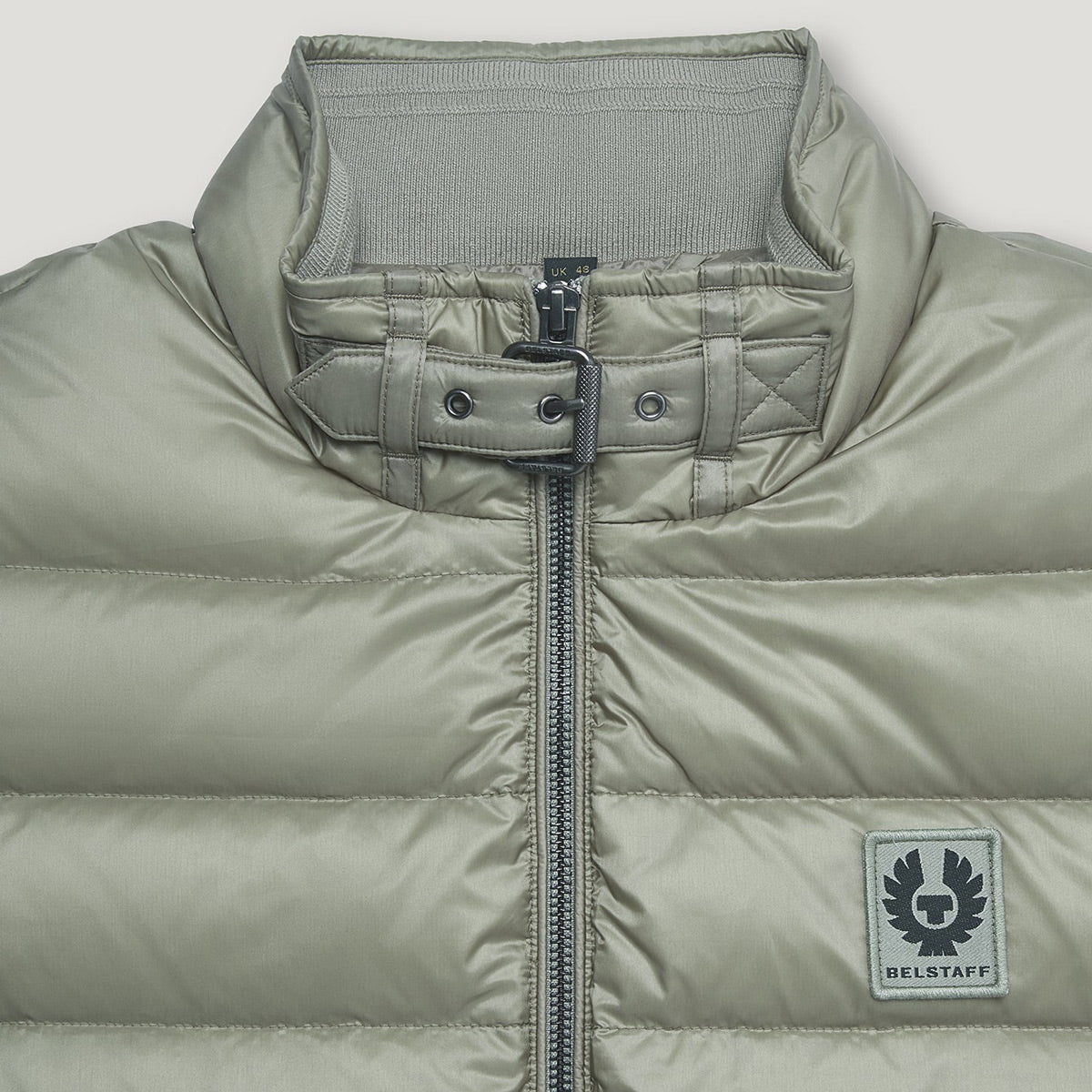 Close-up of a puffer jacket in a muted sage green color, featuring a high collar with a buttoned strap detail, a front zipper, and a square Belstaff logo patch on the lower right side. Belstaff Gilet. Sakks Menswear Southampton.