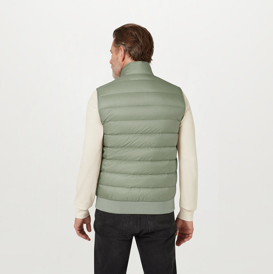 A man viewed from the back wearing a sage green puffer vest over a cream long-sleeve shirt and dark jeans, against a plain light gray studio background. Belstaff Gilet. Sakks Menswear Southampton.