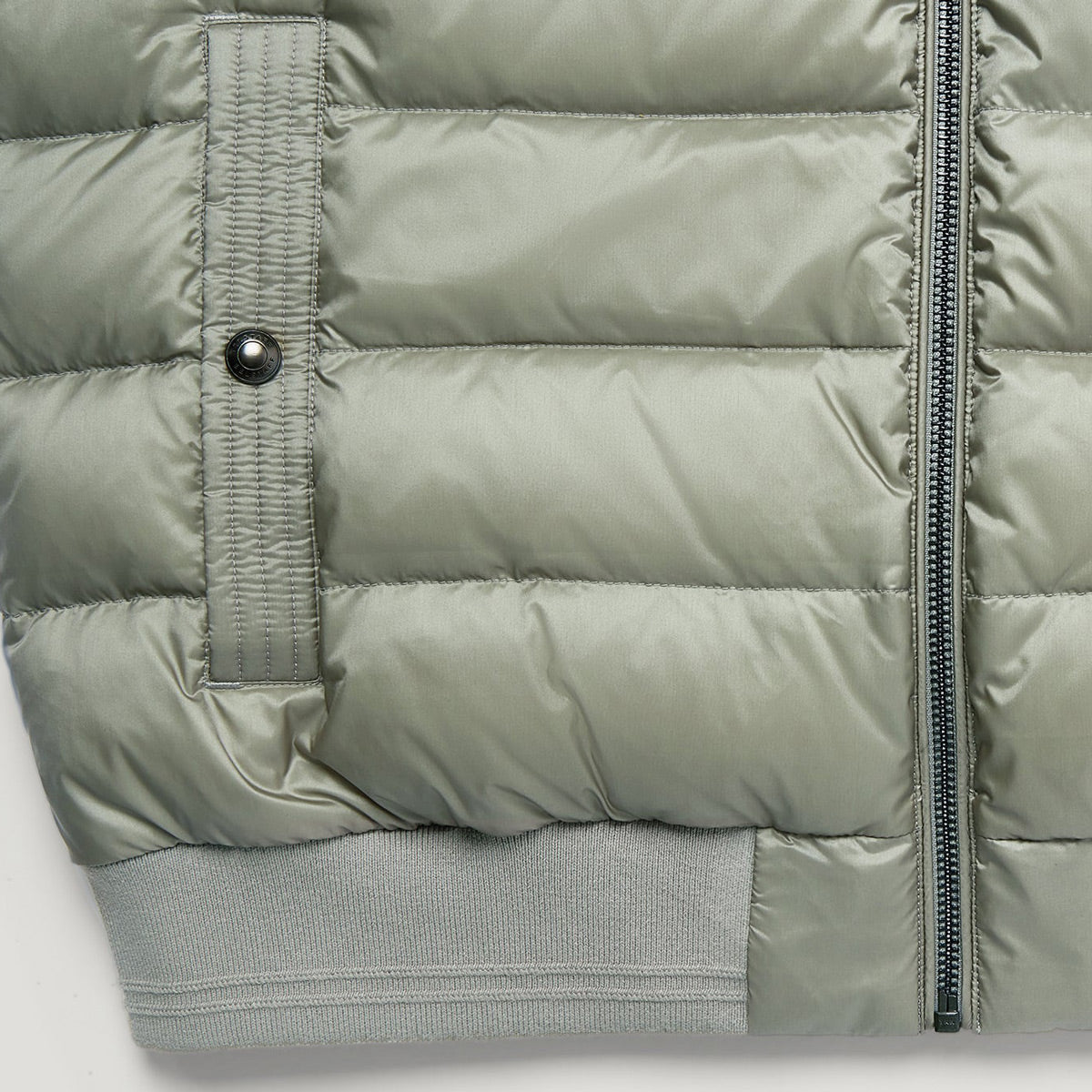 Close-up of a light sage green puffer jacket showing horizontal quilting, a vertical zippered pocket with a snap tab, a black zipper, and ribbed knit cuffs. Belstaff Gilet. Sakks Menswear Southampton.