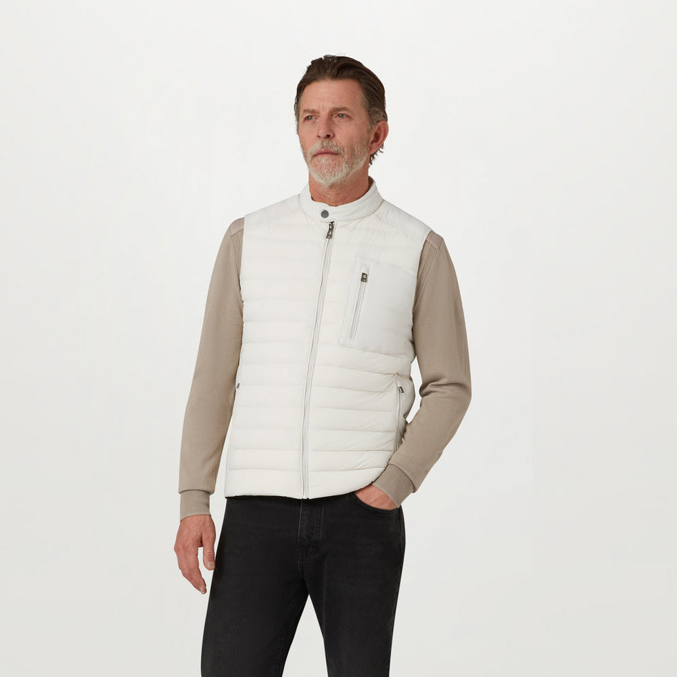 A man wearing a white puffer vest over a beige long-sleeve shirt and black pants, standing against a plain white studio background. Belstaff Gilet. Sakks Menswear Southampton.