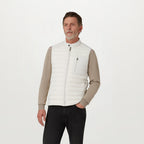 A man wearing a white puffer vest over a beige long-sleeve shirt and black pants, standing against a plain white studio background. Belstaff Gilet. Sakks Menswear Southampton.