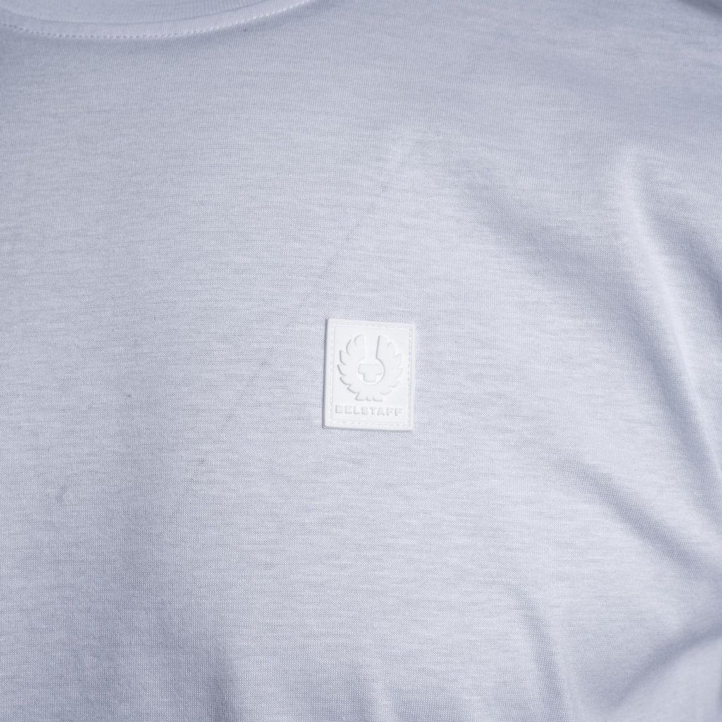 Close-up of a light gray heather crewneck t-shirt featuring a square white embroidered patch on the chest with a winged emblem and the text 'BELSTAFF'. Belstaff T-Shirt. Sakks Menswear Southampton.