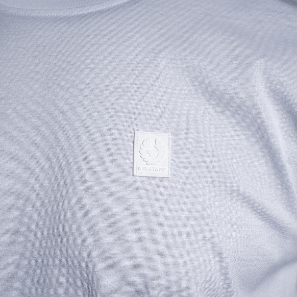 Close-up of a light gray heather crewneck t-shirt featuring a square white embroidered patch on the chest with a winged emblem and the text 'BELSTAFF'. Belstaff T-Shirt. Sakks Menswear Southampton.