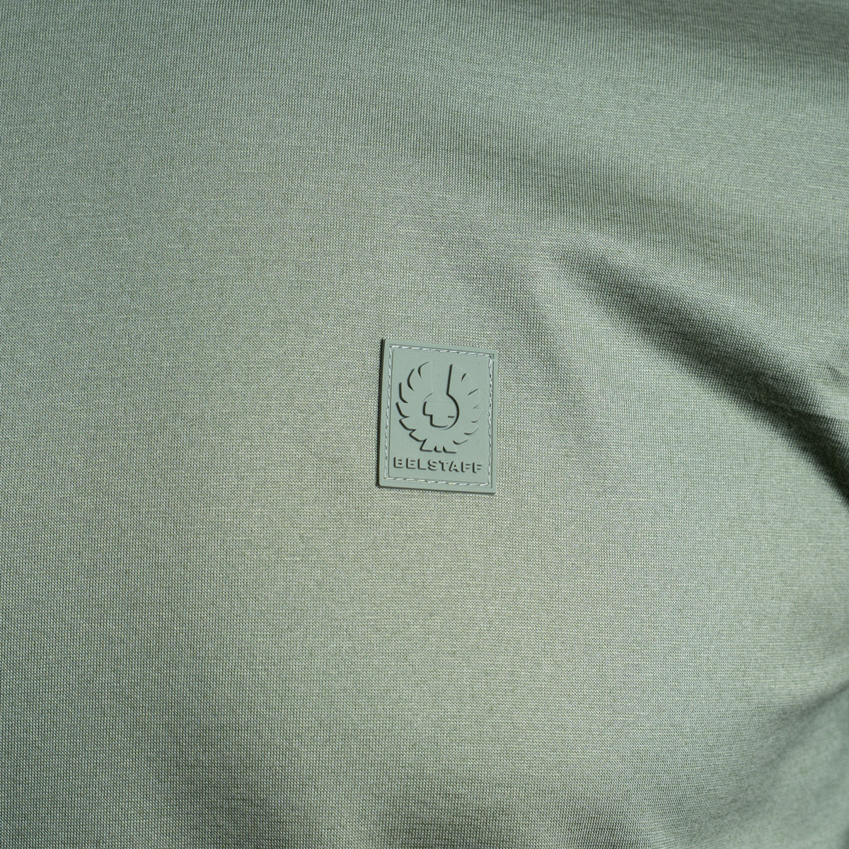 Close-up of a light sage green knit fabric with a small square brand patch reading BULLEYEUF near the center. Belstaff T-Shirt. Sakks Menswear Southampton.