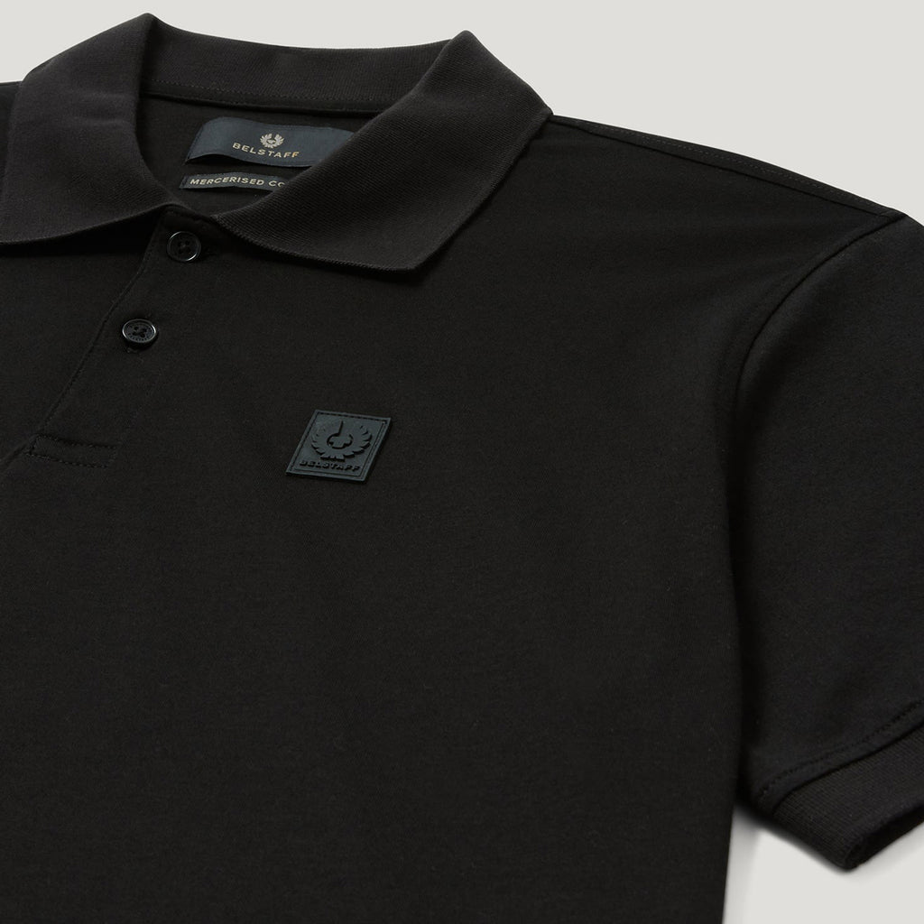 Close-up of a black short-sleeve polo shirt with a collar, two-button placket, and a small square chest patch logo on a light gray background. Belstaff Steel Polo. Sakks Menswear Southampton.