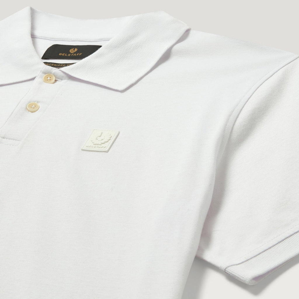 A close-up of a white short-sleeve polo shirt by Fred Perry, featuring a classic collar, two gold buttons, a black brand label at the inner neck, and a small white embroidered logo patch on the chest. Belstaff Polo. Sakks Menswear Southampton.