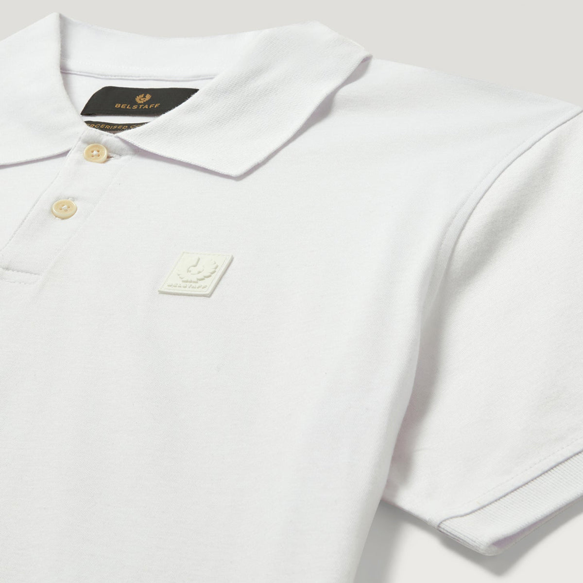 A close-up of a white short-sleeve polo shirt by Fred Perry, featuring a classic collar, two gold buttons, a black brand label at the inner neck, and a small white embroidered logo patch on the chest. Belstaff Polo. Sakks Menswear Southampton.