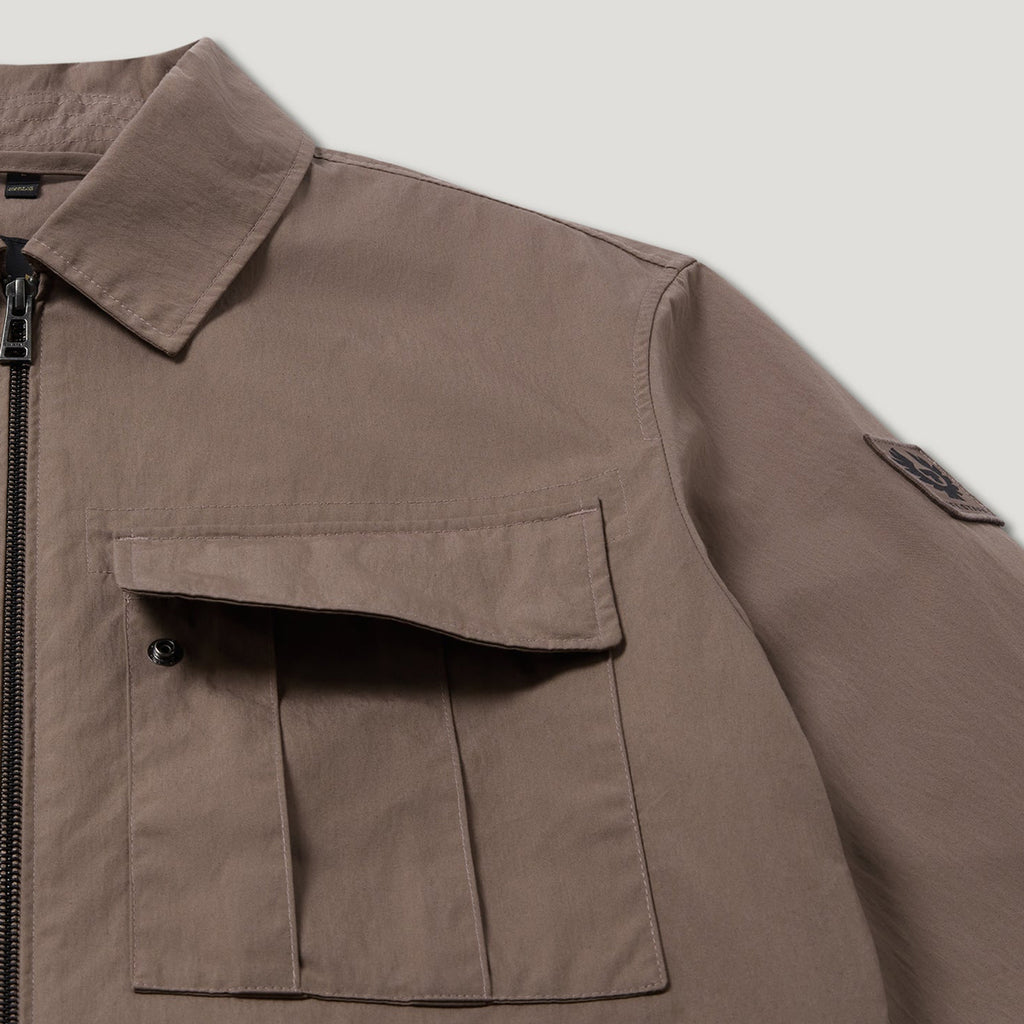 Close-up of a brown utility jacket featuring a pointed collar, a front zipper, a large chest pocket with a buttoned flap, and a small sleeve patch. Belstaff Jacket. Sakks Menswear Southampton.