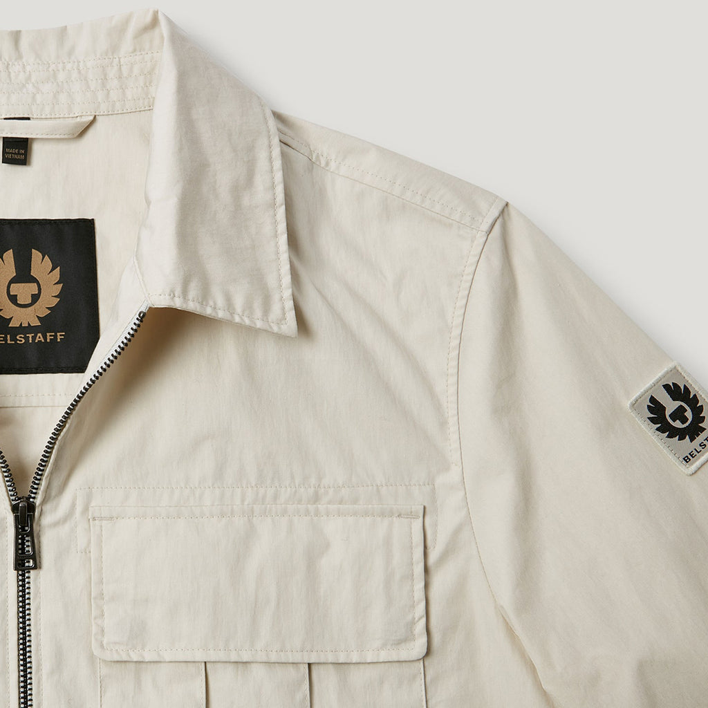 Close-up of a light beige Belstaff overshirt jacket showing the collar, front zipper, chest pocket, and Belstaff logo patches on the inner neck and sleeve. Belstaff Jacket. Sakks Menswear Southampton.