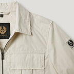 Close-up of a light beige Belstaff overshirt jacket showing the collar, front zipper, chest pocket, and Belstaff logo patches on the inner neck and sleeve. Belstaff Jacket. Sakks Menswear Southampton.