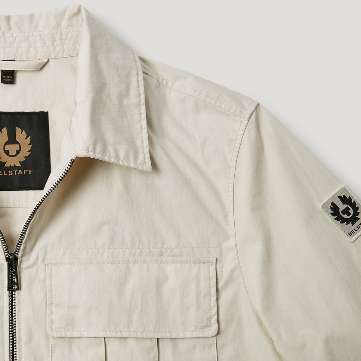 Close-up of a light beige Belstaff overshirt jacket showing the collar, front zipper, chest pocket, and Belstaff logo patches on the inner neck and sleeve. Belstaff Jacket. Sakks Menswear Southampton.
