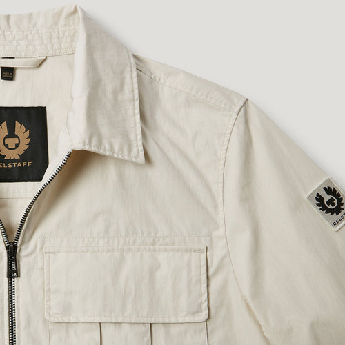 Close-up of a light beige Belstaff overshirt jacket showing the collar, front zipper, chest pocket, and Belstaff logo patches on the inner neck and sleeve. Belstaff Jacket. Sakks Menswear Southampton.