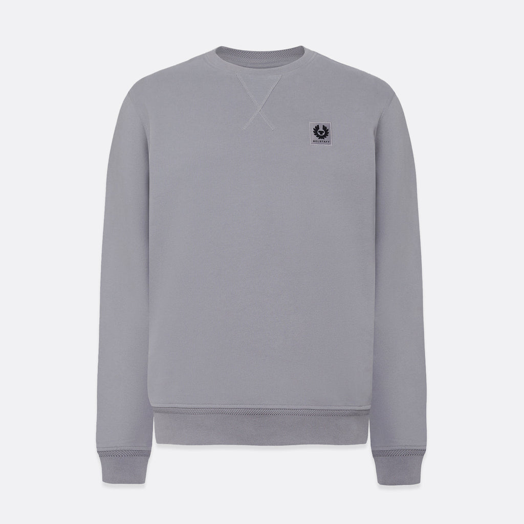 A light gray crewneck sweatshirt with a small black embroidered logo on the left chest, displayed on a clean light gray background. Belstaff Sweatshirt. Sakks Menswear Southampton.