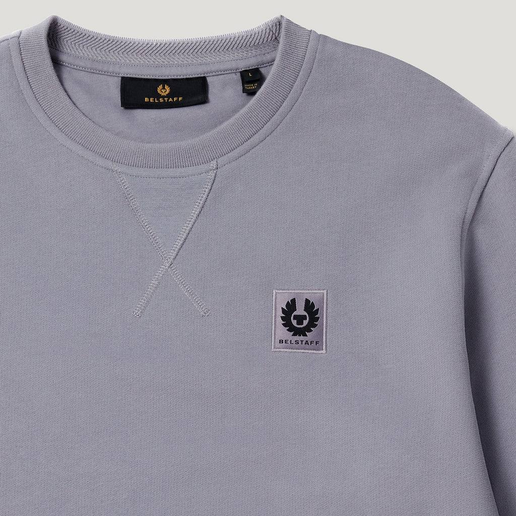 A close-up of a light gray crewneck sweatshirt by Bellruff, featuring a small black and white embroidered logo patch on the left chest and a black neck label. Belstaff Sweatshirt. Sakks Menswear Southampton.