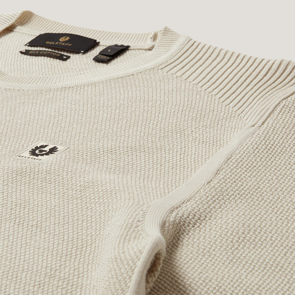 Close-up of a cream-colored ribbed knit sweater with a small embroidered logo on the chest and a black neck label. Belstaff Sweatshirt. Sakks Menswear Southampton.