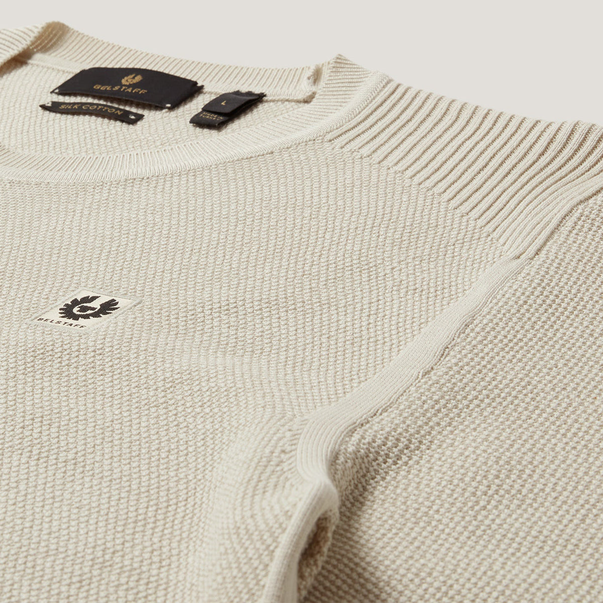 Close-up of a cream-colored ribbed knit sweater with a small embroidered logo on the chest and a black neck label. Belstaff Sweatshirt. Sakks Menswear Southampton.