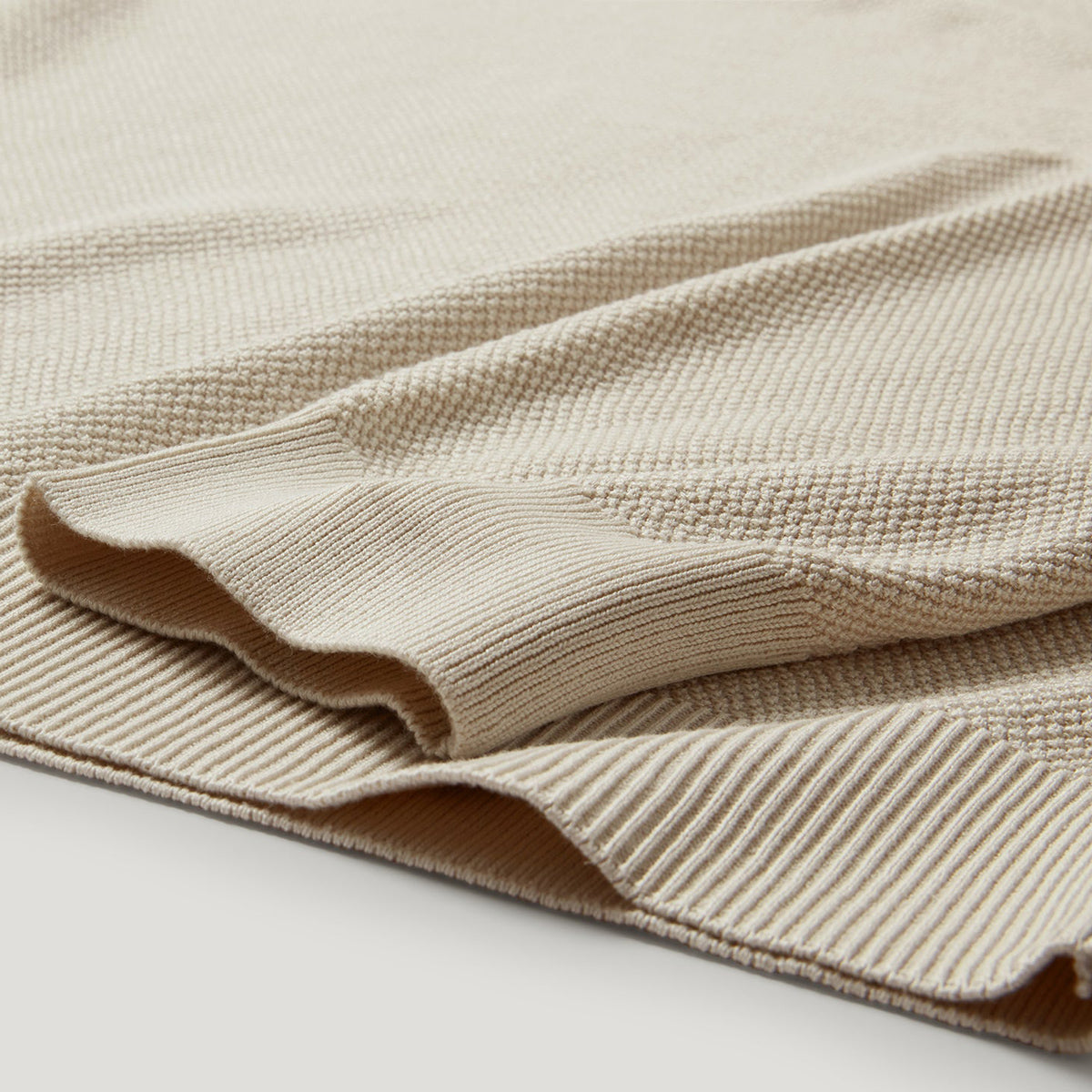 A close-up of a folded beige knit fabric with a textured waffle pattern and ribbed edges, shown on a light neutral background. Belstaff Sweatshirt. Sakks Menswear Southampton.