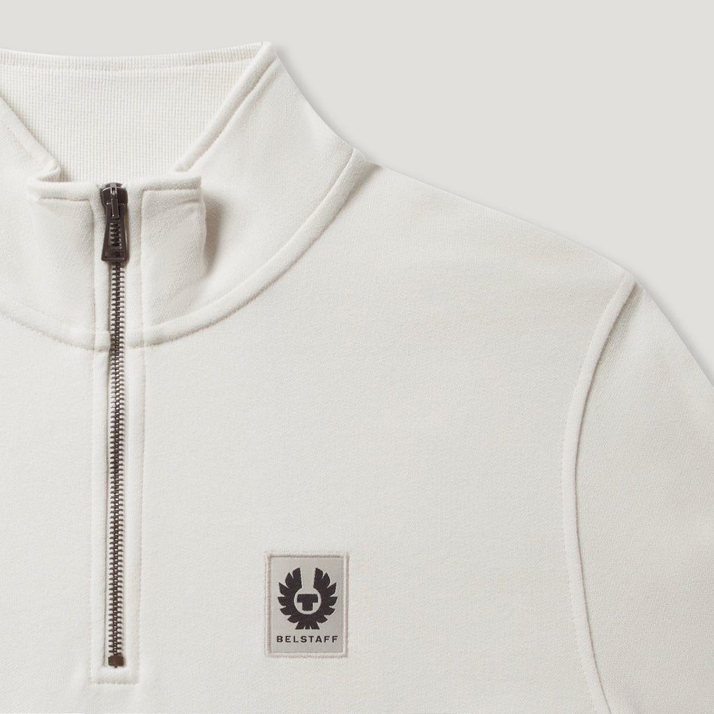Close-up of a white quarter-zip pullover sweatshirt with a black zipper and a Belstaff logo patch on the chest. Belstaff Quarter Zip. Sakks Menswear Southampton.