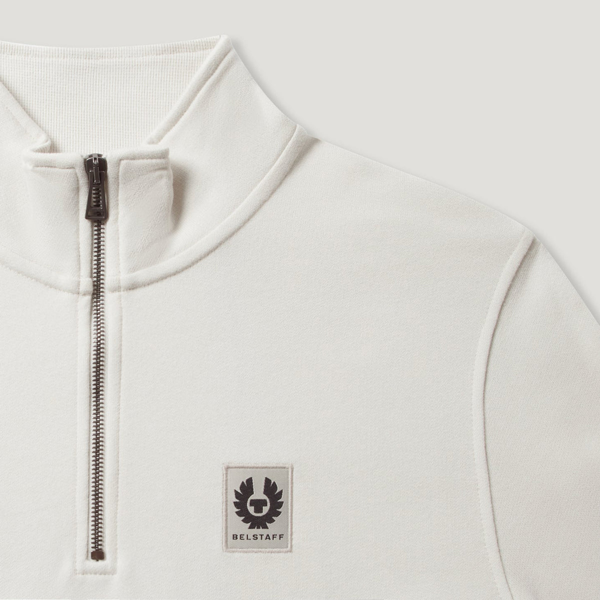 Close-up of a white quarter-zip pullover sweatshirt with a black zipper and a Belstaff logo patch on the chest. Belstaff Quarter Zip. Sakks Menswear Southampton.