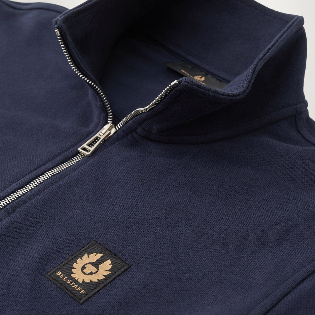 Close-up of a navy blue quarter-zip fleece jacket with a silver zipper and a black embroidered BELL STAFF logo patch on the chest. Belstaff Quarter Zip. Sakks Menswear Southampton.