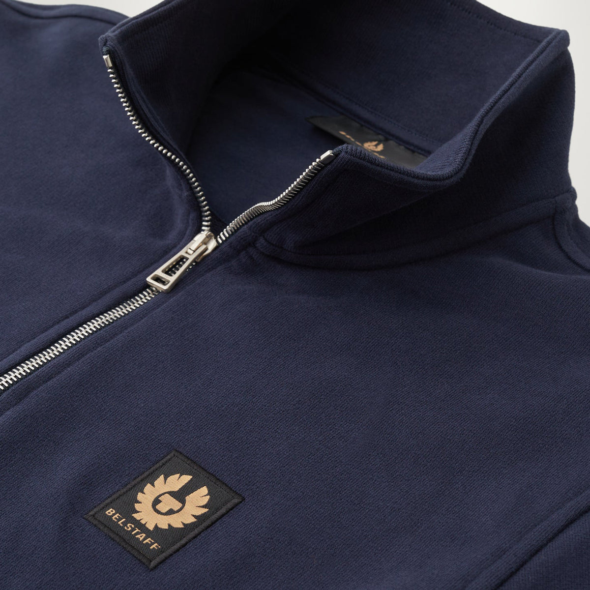 Close-up of a navy blue quarter-zip fleece jacket with a silver zipper and a black embroidered BELL STAFF logo patch on the chest. Belstaff Quarter Zip. Sakks Menswear Southampton.