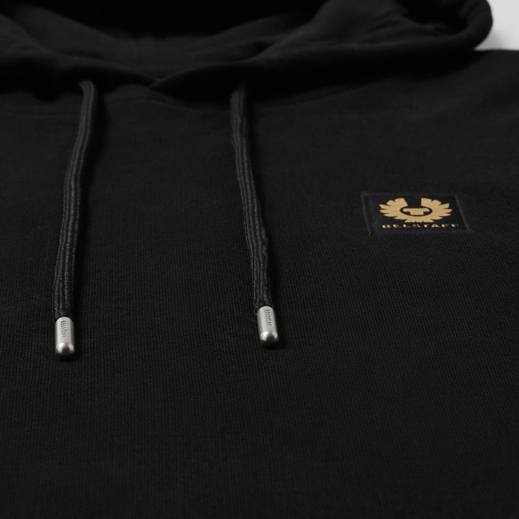 Close-up of a black pullover hoodie with black drawstrings, silver tips, and a small gold chest patch featuring a bird logo and the text 'BELSTAFF'. Belstaff Hoodie. Sakks Menswear Southampton.