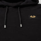 Close-up of a black pullover hoodie with black drawstrings, silver tips, and a small gold chest patch featuring a bird logo and the text 'BELSTAFF'. Belstaff Hoodie. Sakks Menswear Southampton.