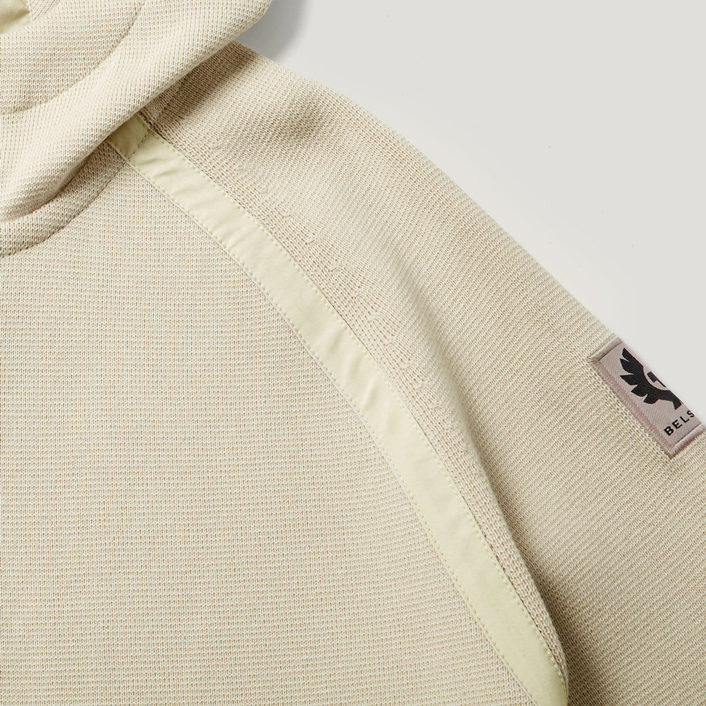 Close-up of a hooded sweatshirt in a light beige textured fabric, showing a pale yellow-green contrast stitched panel and a small black BELLA logo patch on the sleeve. Belstaff Hoodie. Sakks Menswear Southampton.