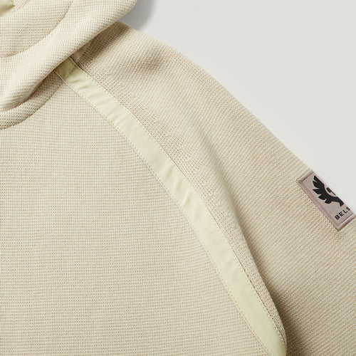 Close-up of a hooded sweatshirt in a light beige textured fabric, showing a pale yellow-green contrast stitched panel and a small black BELLA logo patch on the sleeve. Belstaff Hoodie. Sakks Menswear Southampton.