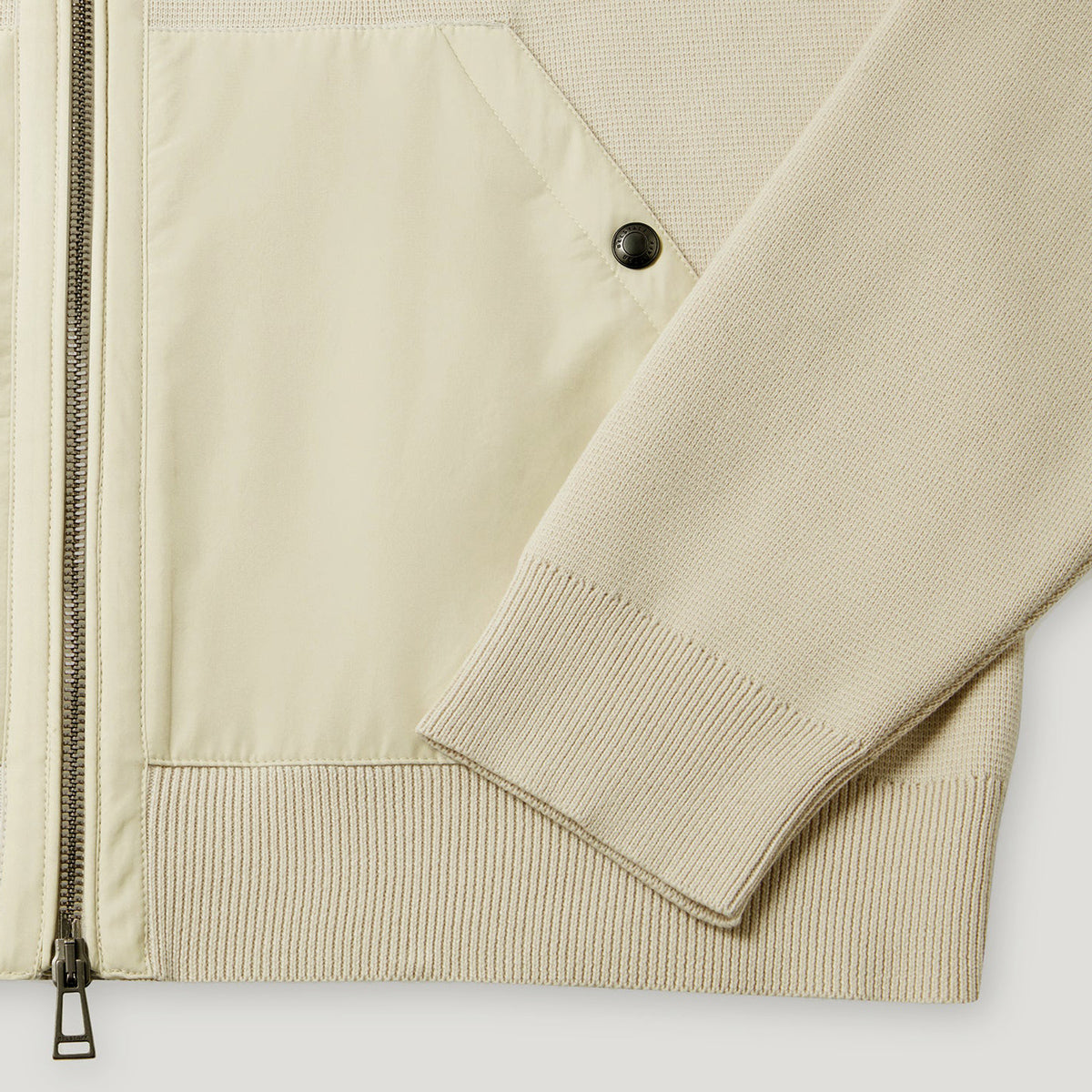 Close-up of a light beige jacket showing a smooth zippered panel, a ribbed knit sleeve, and a black snap button detail. Belstaff Hoodie. Sakks Menswear Southampton.